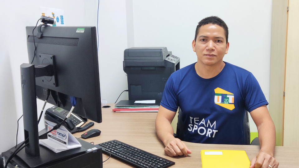Foto colorida com Dorisvan centado em uma mesa que tem um computador e um caderno amarelo a sua frente. 